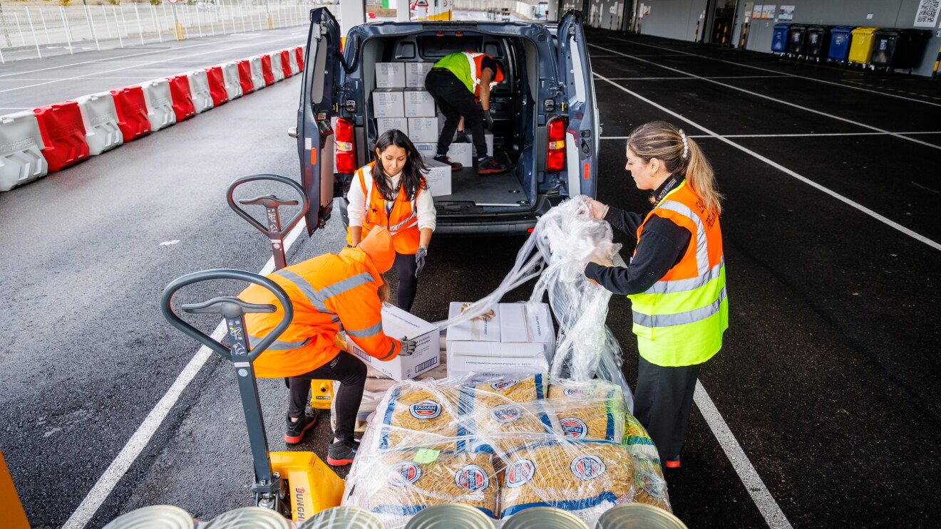 En primer plano latas de comida y paquetes de macarrones. En el margen izquierdo se ven dos toros semiautomáticos. Tres mujeres quitando el plástico transparente que envuelve los palés. Dos de ellas van con chaleco naranja y jersey debajo y la tercera lleva un abrigo de color naranja. En el fondo se ve una furgoneta con el maletero abierto y semilleno. El hombre va con chaleco, guantes y manga corta.