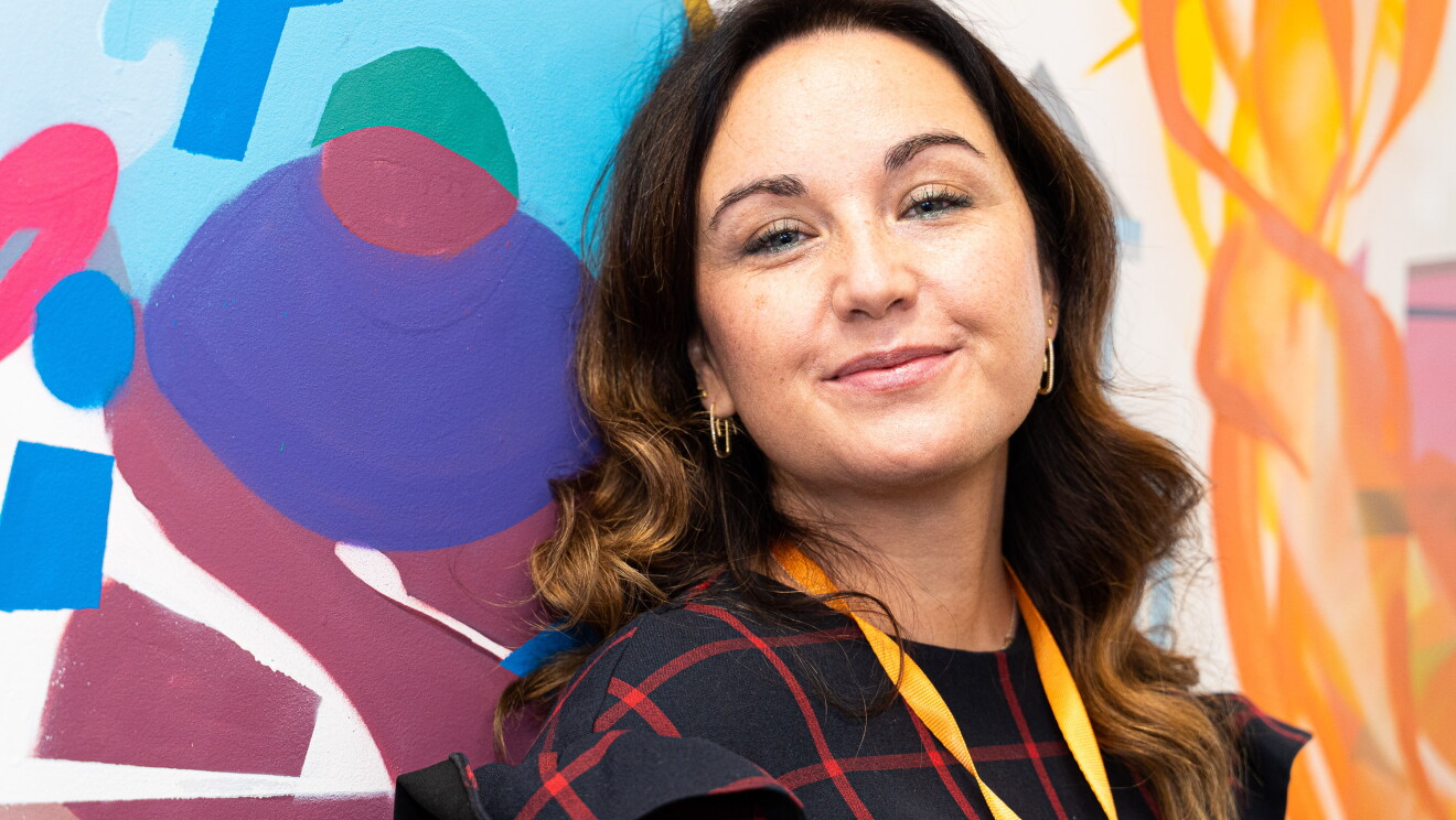 Elena Bris trabajando en las oficinas de Amazon en Madrid.  Elena Bris trabaja en un proyecto regulatorio de mercancías peligrosas. Mirando a cámara en primer plano. De fondo una prared estampada con pinceladas de difernetes colores. Elena lleva un bagde con una cinta de color amarillo y un vestido azul con cuadros de color rojo. Tiene el pelo largo y ondulador. Lleva pendientes y los ojos pintados de azull. Lleva pendientes. 