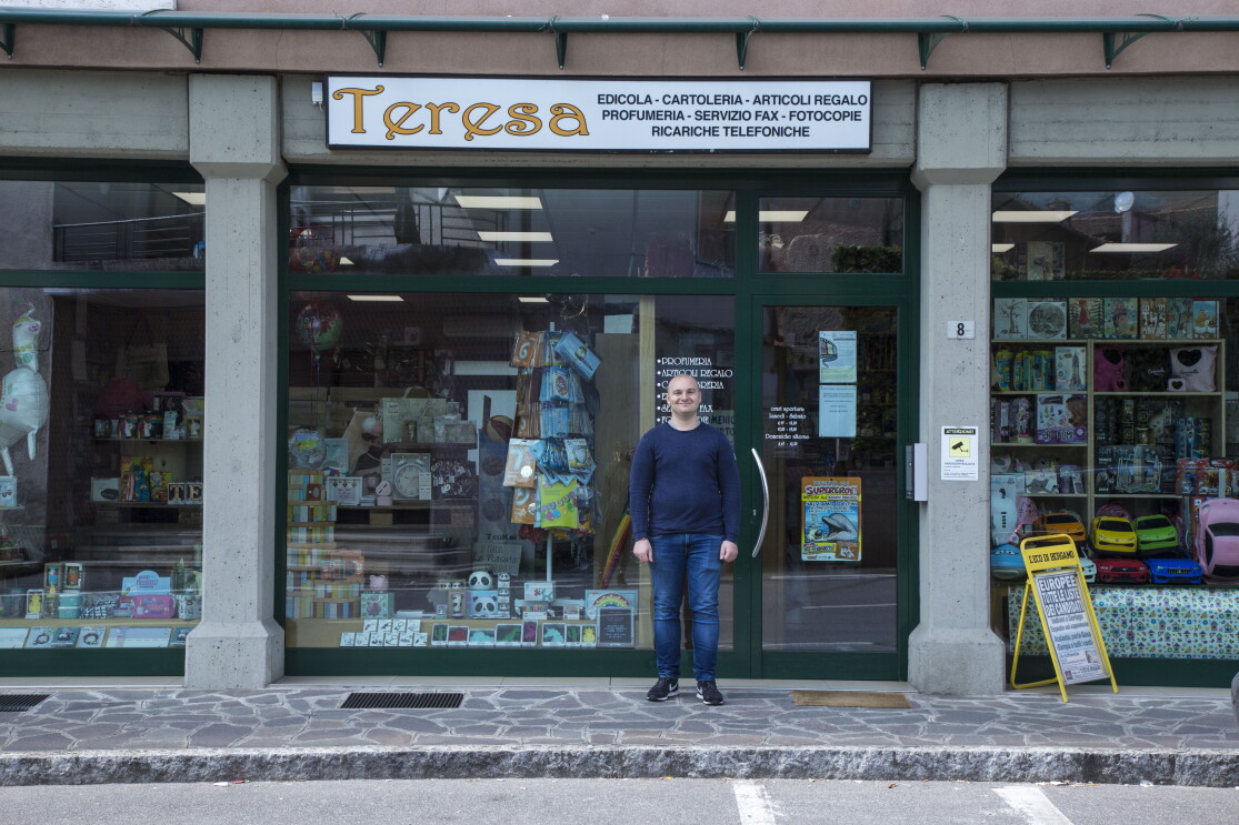 Andrea Locatelli posa di fronte all'ingresso del negozio di Teresa. L'insegna dice "Teresa - edicola, cartoleria, articoli regalo, profumeria, servizio fax, fotocopie, ricariche telefoniche."