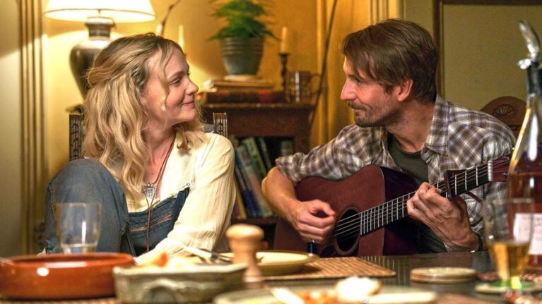 Carey Mulligan and Tom Basden sit at a dinner table, looking at each other as Basden plays a guitar