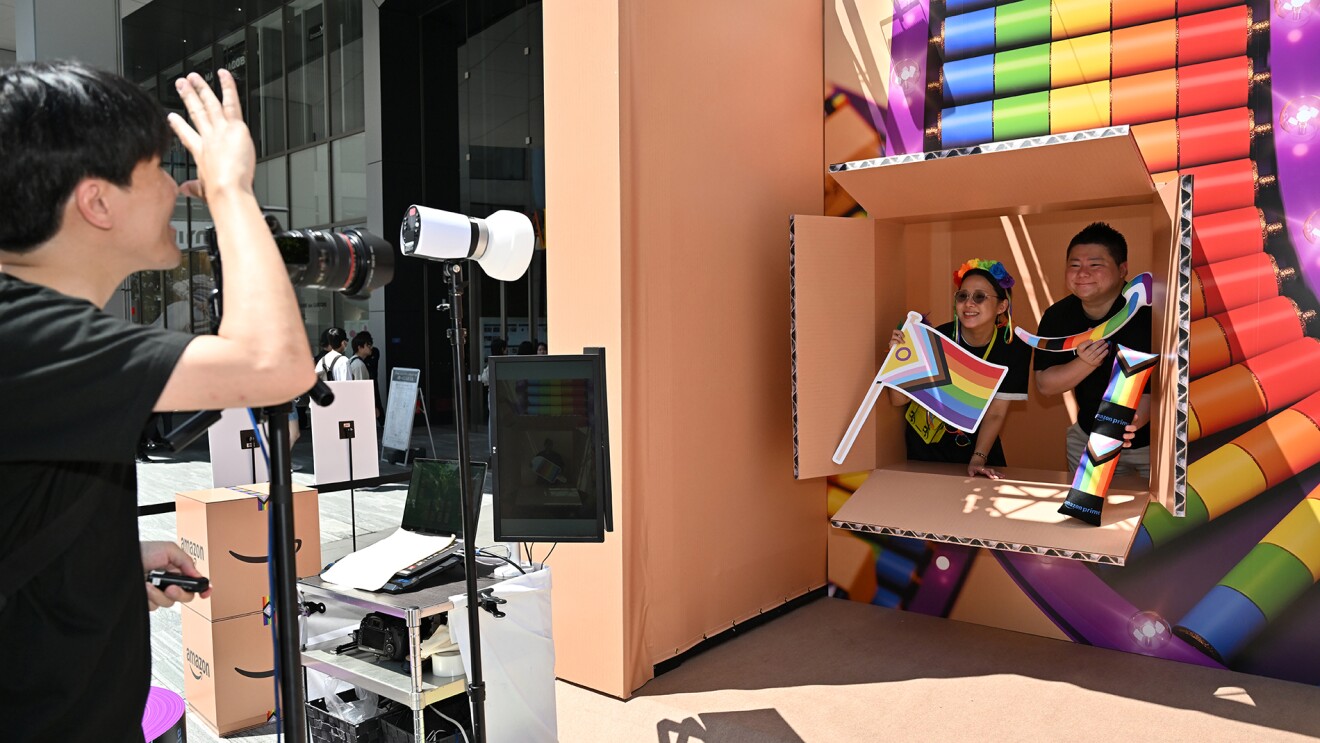 段ボールが開いた形のフォトブースの中でレインボーカラーのパネルを持ってポーズをとっている人たちと、その人たちに手で合図をしているカメラマン