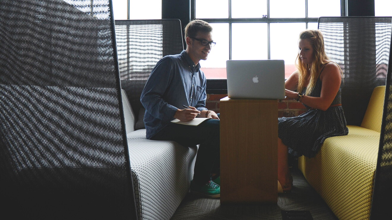 Man taking notes as his female colleague works on a laptop, in a casual work space.