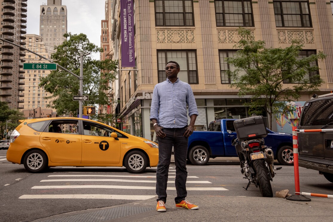 An image of a man standing on the sidewalk in the middle of a large city. There are cars and a taxi cab on the street behind him.