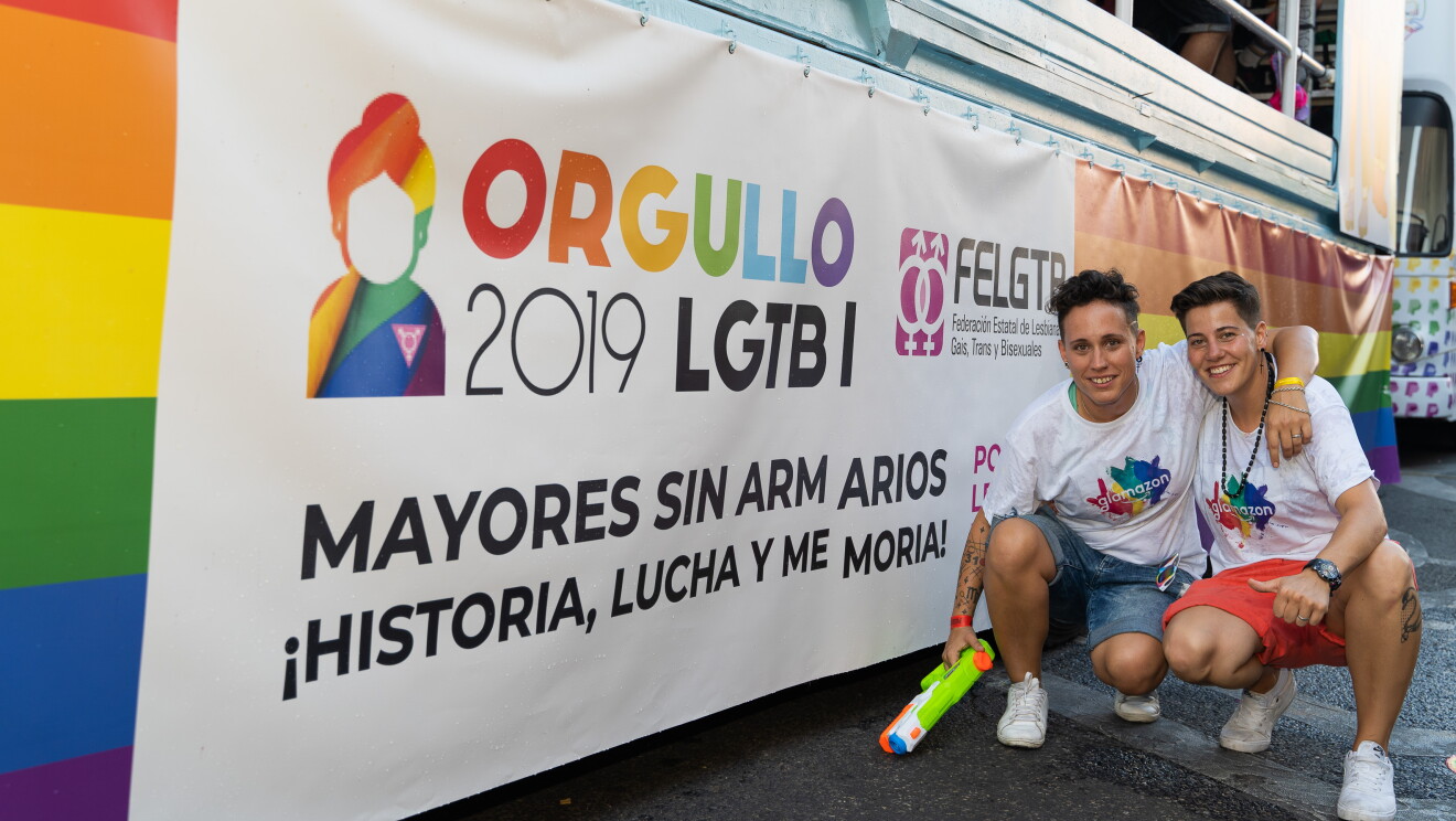 María García y Susana Torres delante del cartel de la carroza de glamazon del desfile del Día internacional del orgullo LGTBI en Madrid. La carroza tiene la bandera del arcoiris y encima el cartel oficial del desfile con el mensaje Mayores sin armarios. ¡Historia, lucha y memoria! María y Susana se encuentran agachadas delante del cartel. María lleva una pistola de agua en la mano derecha y con la izquierda abraza por el cuello a Susana. Las dos llevan la camiseta de glamazon y van en pantalón corto y deportivas. 