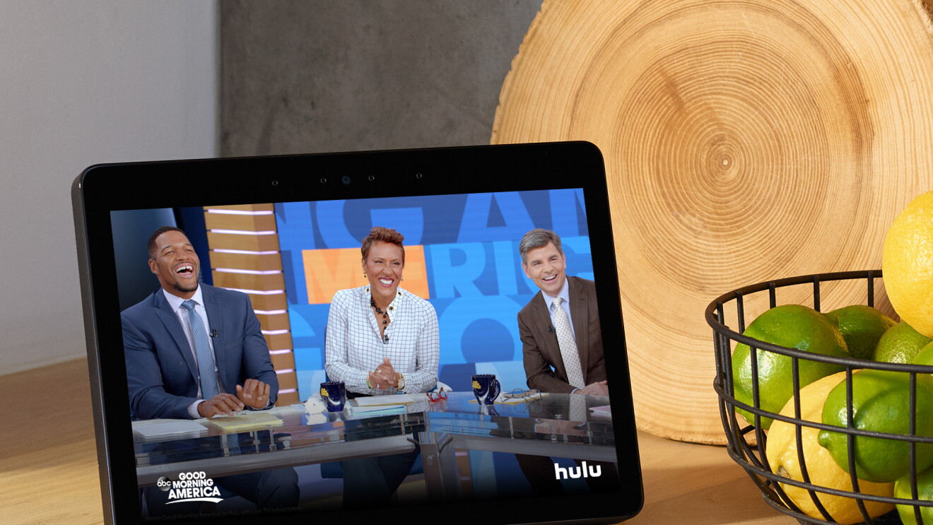 An Echo Show device on a kitchen counter, with Good Morning America displayed on the screen. To the right of the device is a cutting board made of a slice of wood, and a metal basket filled with lemons and limes. 