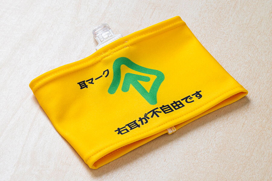 白に近い木目調の平らな表面に置かれた黄色い腕章。上辺に透明な留め具が付いている。腕章の上の方に「耳マーク」の黒文字とその右側に緑の線で描かれたマーク。耳マークは線で囲まれた上向き矢印が耳の形に見える。下部に「右耳が不自由です」の黒文字。