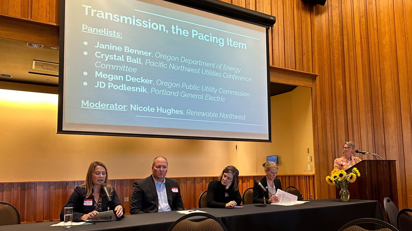 Four panelists sitting at a table and a moderator standing at a podium at the Oregon Clean Grid Summit.