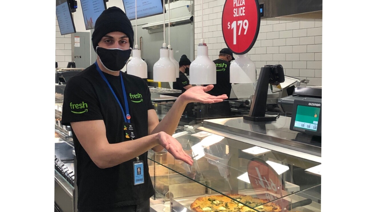 Rotem stands with her hands up to display the fresh and hot pizza by the slices for sale. She wears a black beanie, a black Amazon Fresh T-Shirt, and a face mask.