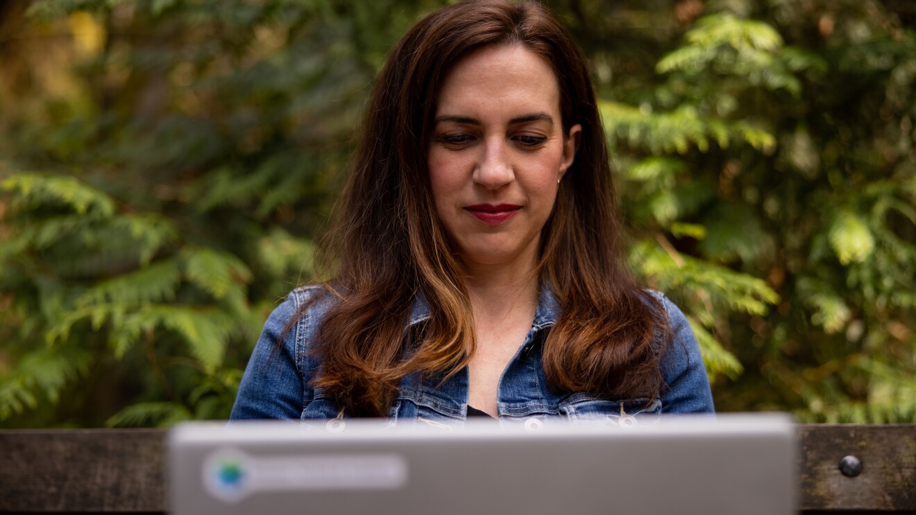 Ashely Rajagopal sits on a bench outside with vibrant greenery behind her. She is focused while she works on her laptop.
