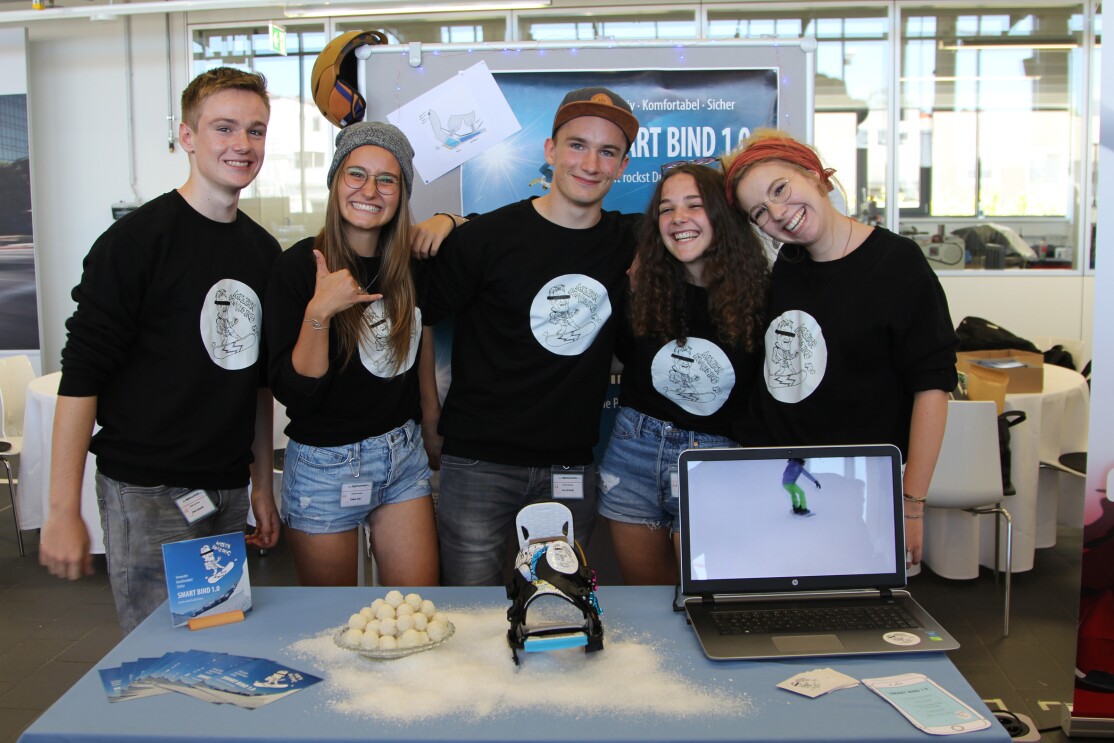 Fünf Jugendliche mit schwarzen Shirts, Aufdruck "Modern Boarding" und Jeans stehen vor einem Tisch. auf ihm stehen ein Laptop, eine Snowboardbindung. Zusätzlich sind ein halbes Dutzend Flyer zu sehen.
