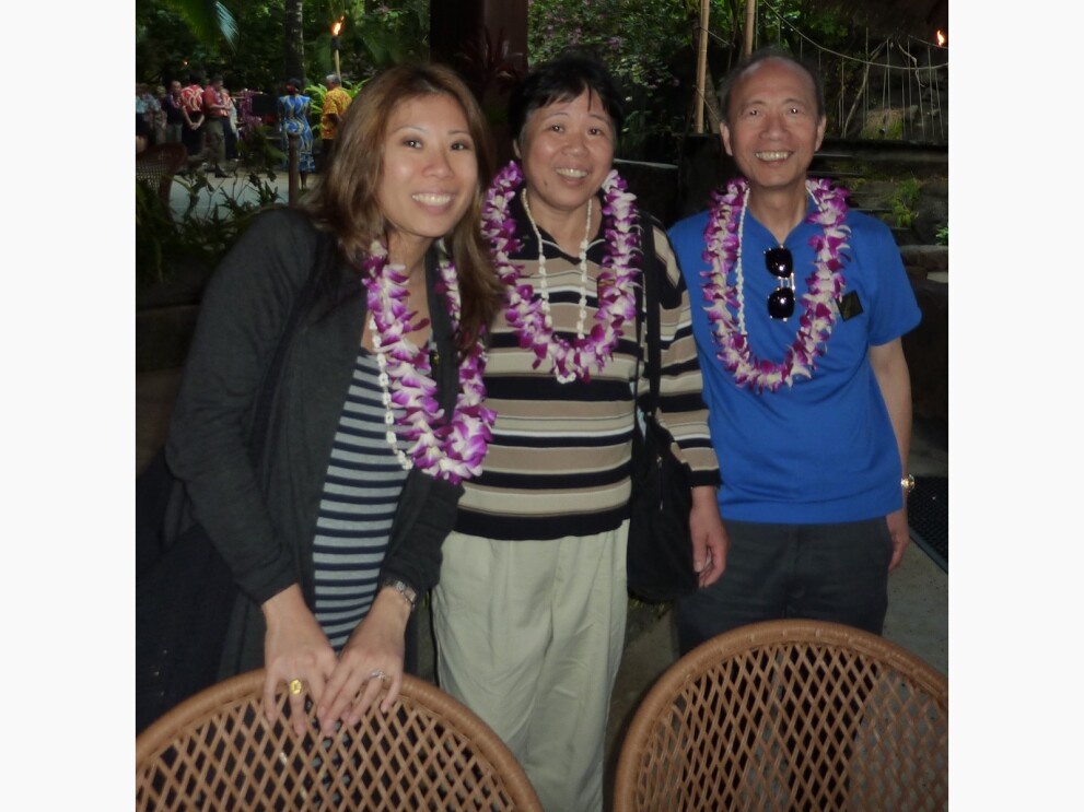 Angela smiling and posing with her parents. They are in tropical setting and wearing flower leis.