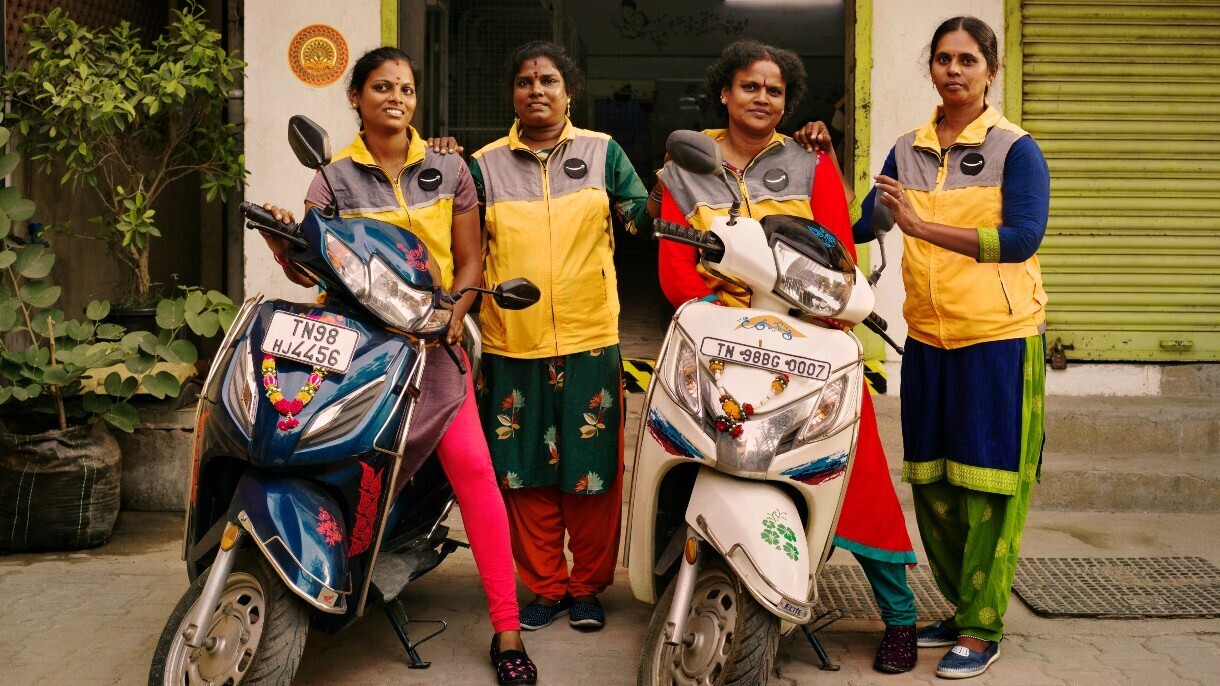 Cuatro mujeres indias vestidas con el chaleco amarillo de Amazon. La primera y la tercera están subidas a dos motos de color azul y blanco, respectivamente. La segunda y la cuarta de pie agarrando el hombro a su compañera. Las cuatro van vestidas con trajes tradicionales. Están en la calle, delante de una estación logística con la persiana subida.  