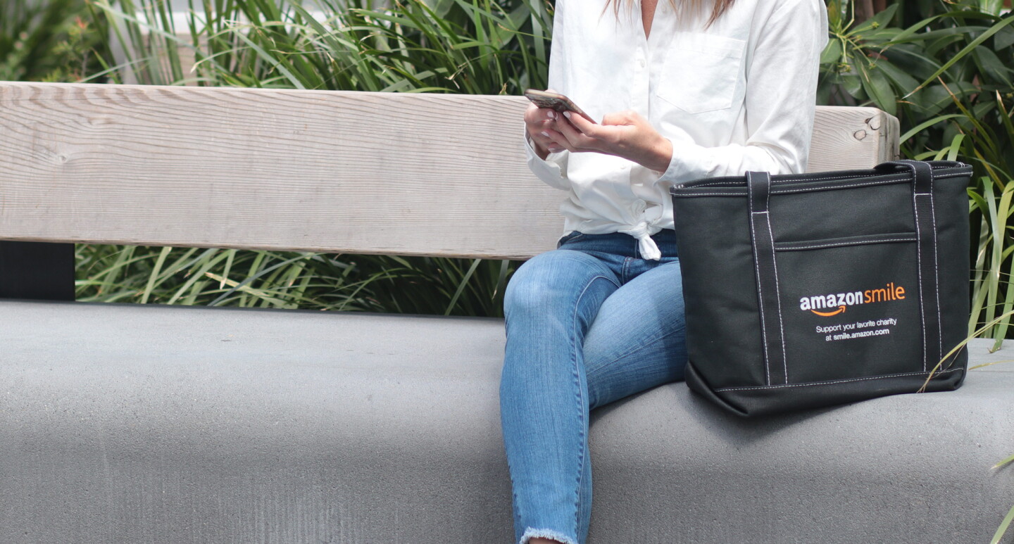 A woman on a bench holds a smartphone. She is seated next to a tote bag with the AmazonSmile logo and the words "Support your favorite charity at smile.amazon.com."
