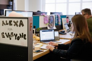 Two people sit at their desks which are side-by-side. There is a white board that reads "Alexa for everyone"