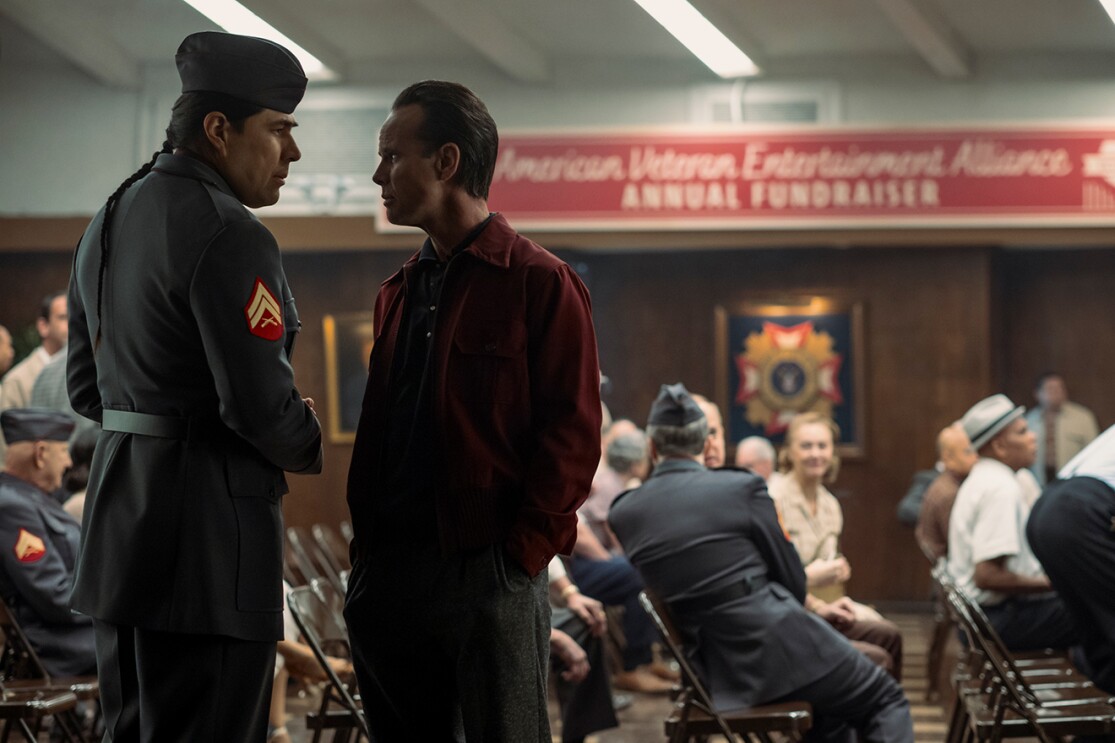 人々が集う集会室で向き合ってにらみ合う2人の男性。左側は濃いグレーの軍服、右側は暗い赤のシャツを着用。2人の背後にはりたたみ椅子が並び、人々が着席している。天井近くに赤い横断幕。