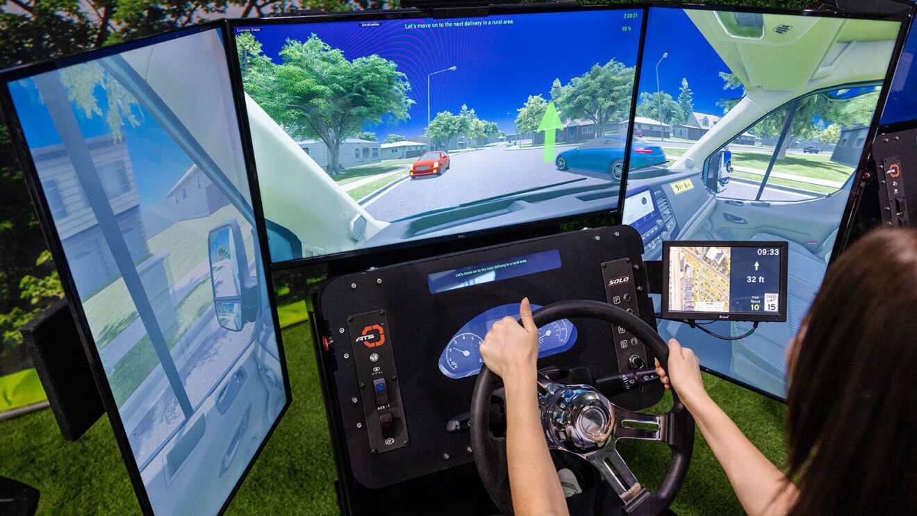 An image of three large screens simulating the driving experience on an Amazon Rivian delivery van. There is a simulated dashboard and steering wheel below the screen, and a woman is holding the steering wheel while sitting in the driver's seat.