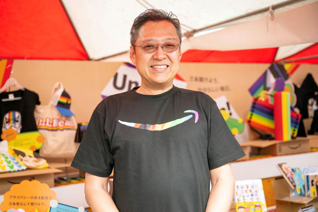 さまざまな商品が並ぶ展示台の前に立つサングラスをした男性。Amazonのロゴが入った黒いTシャツを着ている