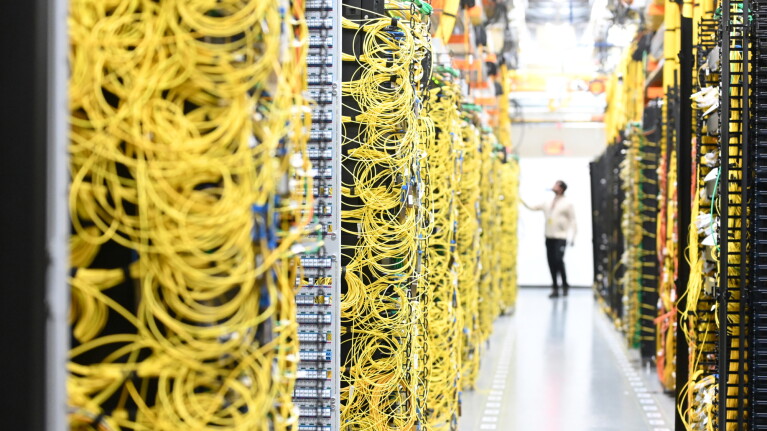A technician stands in the aisle of a data center between towering server racks densely packed with bright yellow and orange network cables.