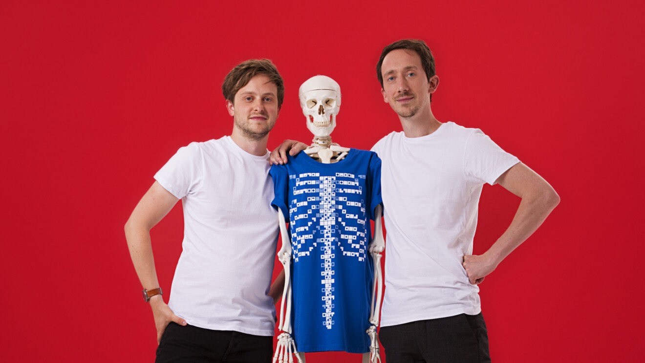 Ed Barton e Ben Kidd, fondatori di Curiscope. I due ragazzi, in t-shirt bianca e pantaloni nero, abbracciano uno scheletro con una t-shirt blu. Sfondo rosso.