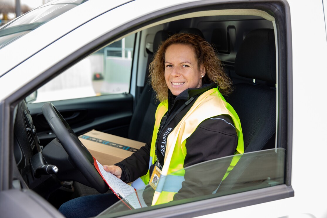 Eine Frau mit halblangen Haaren und Sicherheitsweste in einem Lieferfahrzeug am Steuer.