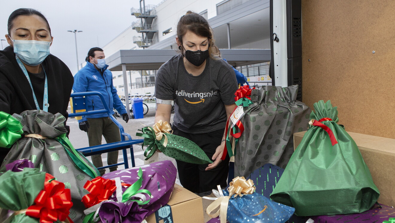 Dos trabajadores de Amazon cargando una furgoneta para realizar la donación a Fundación Acogida (Madrid). Se ve la furgoneta desde dentro y en la puerta trasera hay dos chicas organizando regalos que van en bolsas de diferentes colores: verde, gris, morado y azul. Las dos chicas est´´an situadas en la puerta trasera de la furgoneta. La de la izquierda lleva mascarilla azúl y sudadera negra. La otra chica lleva el pelo recogido, mascarilla negra y camiseta de Amazon gris, manga corta, con la etiqueta DeliveringSmiles. De fondo se ve el conductor con mascarilla azul, chaqueta azul y pantalón gris. Y de fondo el exterior de un centro logístico de Amazon. 
