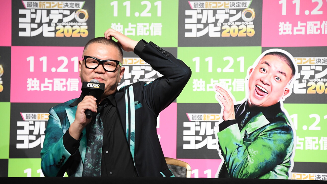 ピンク、黄緑、黒の市松模様の壁を背景に、マイクを持つ黒ぶち眼鏡の男性。黒と緑マーブルのスーツ衣装で、左手で頭をかくポーズ。右側に、同じ衣装の別な男性の等身大パネル。