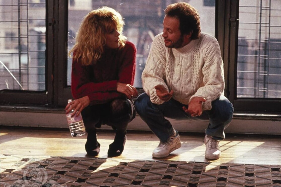 A blonde woman in a red jumper and a man in a white jumper looking at each other in an empty apartment