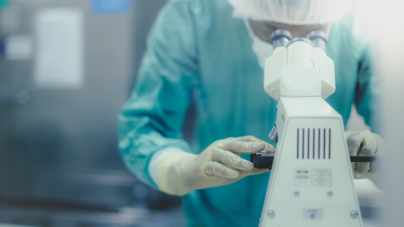 A person in a PPE examines a sample in a microscope