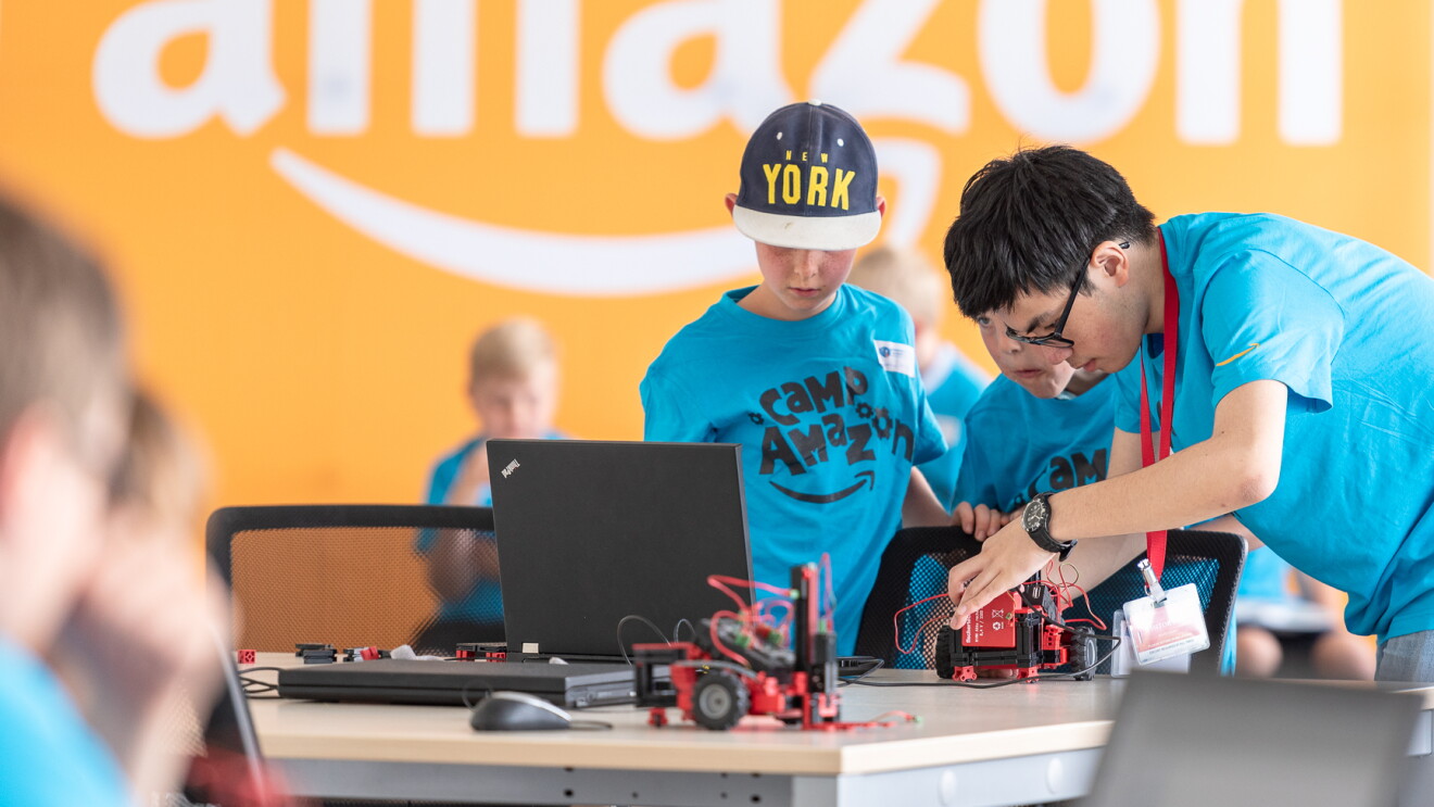 2 Kinder in blauen T-Shirts mit der Aufschrift "Camp Amazon" basteln an einem Tisch an kleinen Gabelstaplern