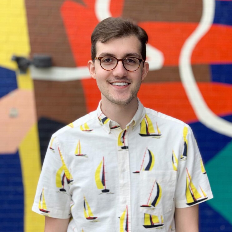 Amazonian stands in front of a colorful wall. He wears a button down with sailboats and glasses.