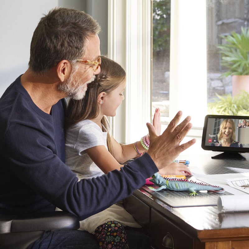 A little girl sits on a man's lap at a desk. They are both waving and looking at an Echo Show device, on which a woman is smiling and engaging with them. 