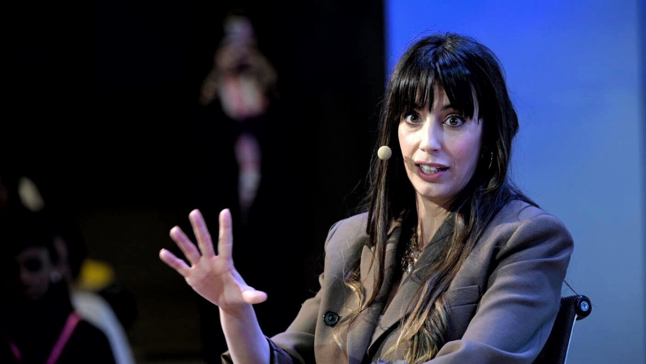 Almudena Cid, exgimnasta olímpica, aparece en primer plano. Está sentada en una silla negra. Lleva un traje chaqueta de color marrón, el pelo negro largo hasta el pecho y un micrófono en la mejilla. Está hablando y tiene las uñas pintadas de rojo.