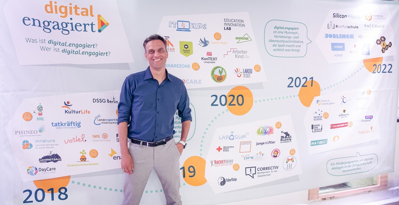 Ein Mann von Amazon (Rocco Bräuniger, Country Manager Amazon DACH) steht vor einem Zeitstrahl, der die Entwicklung der Förderinitiative digital.engagiert abbildet.