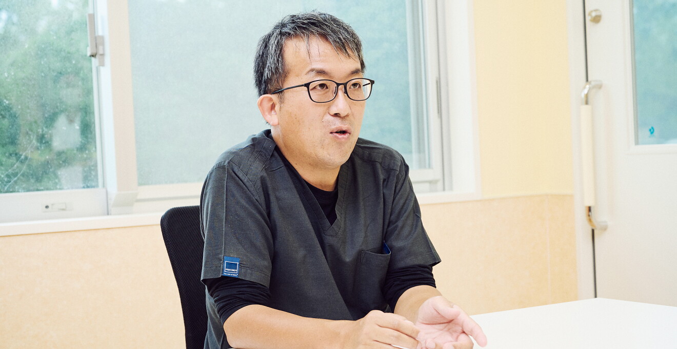病院の会議室で窓を背に座っているメガネをかけた男性が話している。濃いグレーの医師のユニフォームを着ている。