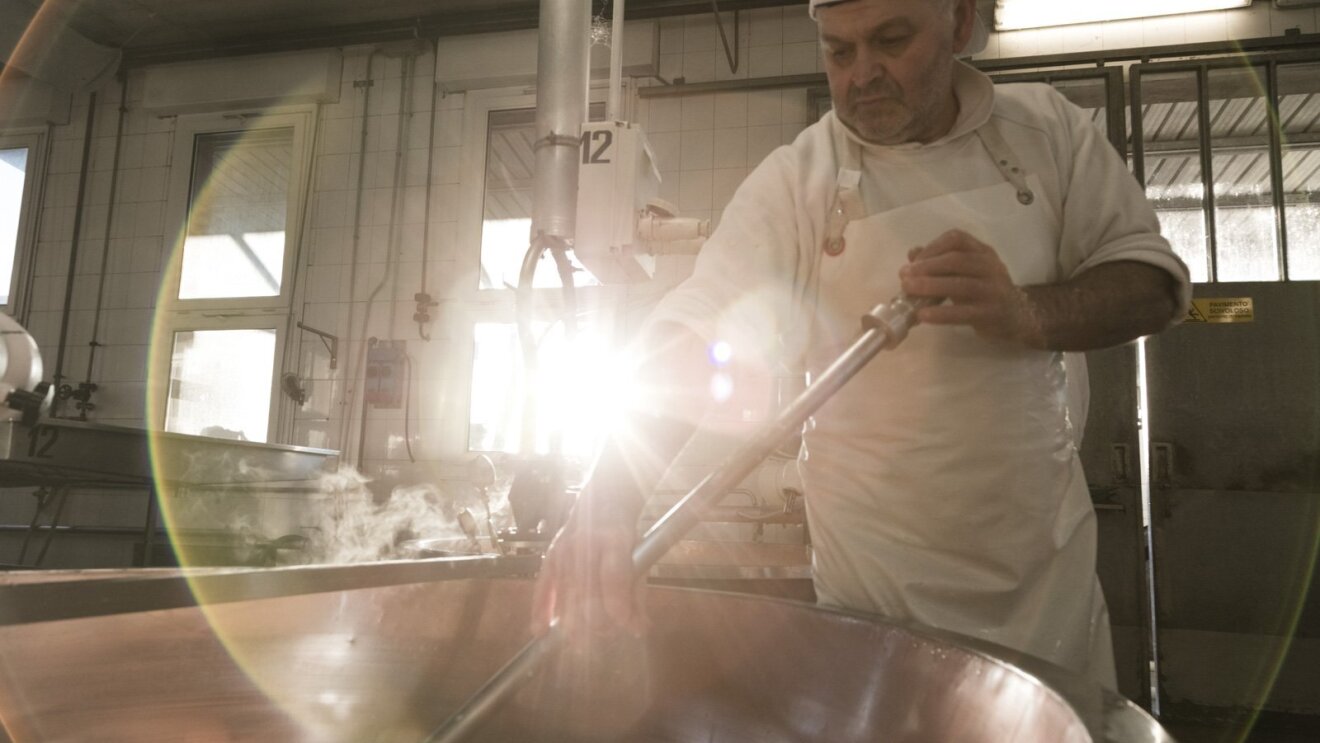 Immagine di un uomo mentre lavora il formaggio nel laboratorio della Latteria Due Madonne. L'uomo regge lo strumento chiamato 'spino' e con questo gira la cagliata dentro un grosso pentolone. L'uomo è vestito di bianco e indossa un grembiule e un cappello. La scena è ripresa di giorno.