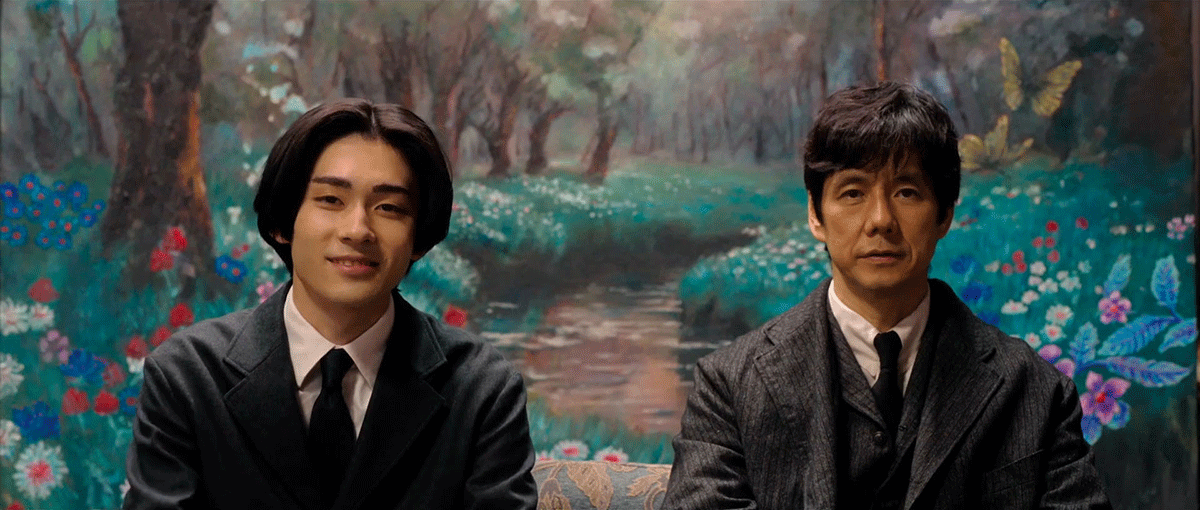 花咲く森の道が奥に続く風景画を背景に、2人の男性の上半身。左側は黒いスーツの若者で、右側は濃いグレーにストライプが入ったスーツの男性。