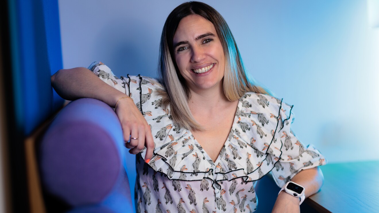 Lara Fajardo va con una camisa blanca estampada, con el pelo hasta el hombro con mechas negras y decoloradas hasta el rubio en las puntas. Con pantalones blancos y un cinturín de color negro. Con uñas rosas y un anilo en cada mano. En la mano derecha un reloj. Esta mano está apoyada en la mesa y la otra en un sofá de color azul y morado.