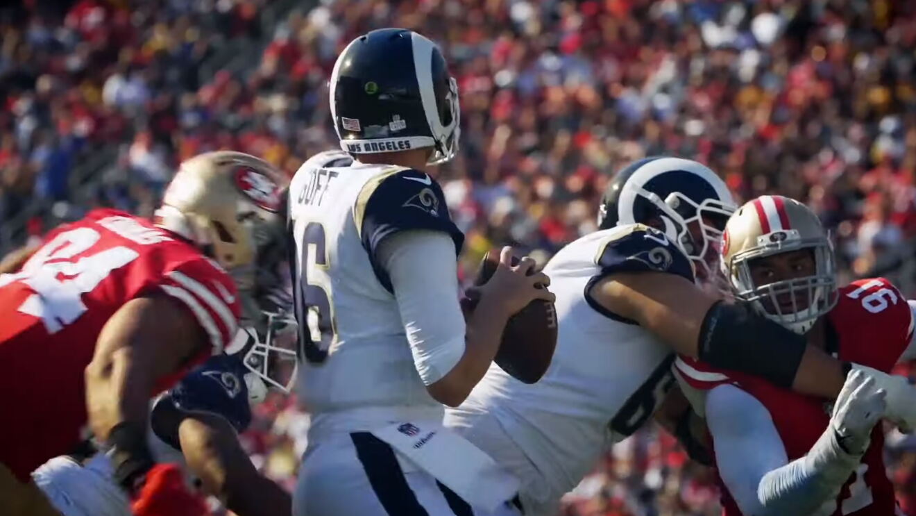 NFL Rams take on the San Francisco 49ers. Quarterback Jared Goff holds the ball as he readies to throw it. A teammate blocks an opponent, while another opponent rushes to make a sack.  