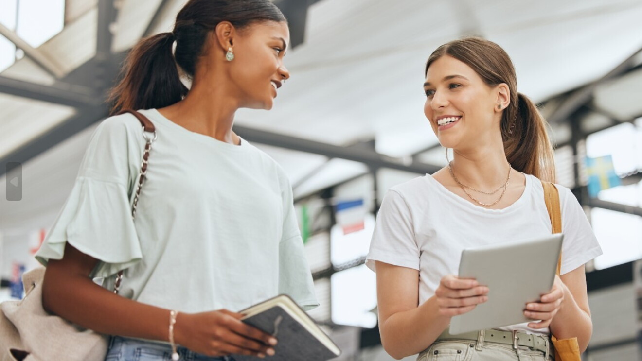 Due ragazze camminano con dei libri in mano e sorridono l'una all'altra. Entrambe indossano una t-shirt bianca e sembrano trovarsi in un corridoio universitario. 