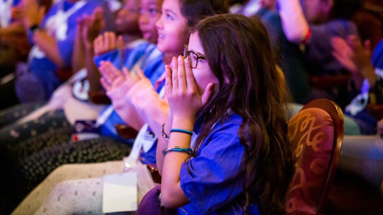 A young girls sits in the audience at Girls' Tech Day with her hands over mouth in excitement.