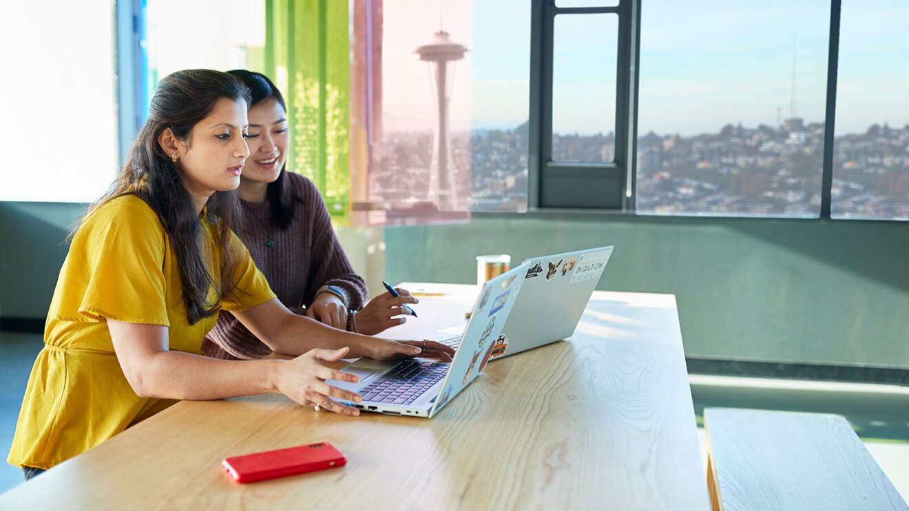 Two women work at a table on their computers at the Amazon office with a view of the Seattle Space Needle.