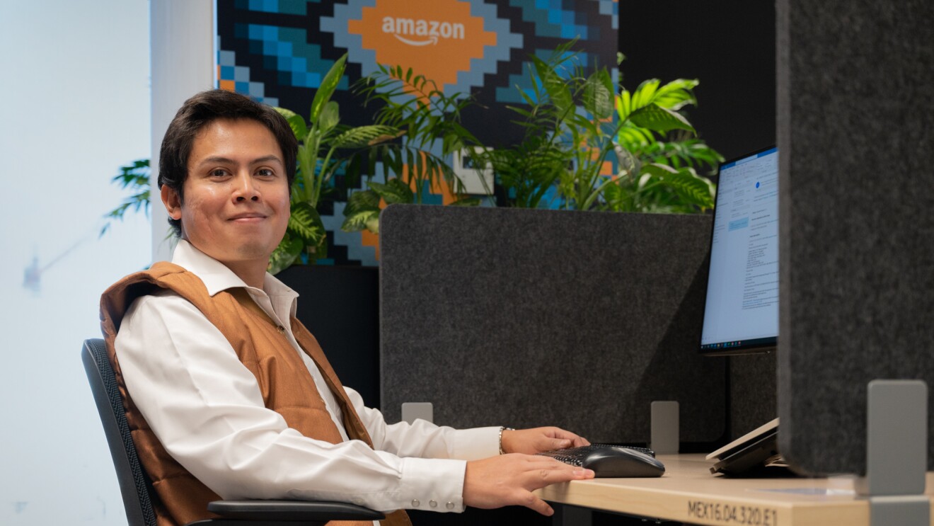 Un trabajador de Amazon en una oficina, sentado en una mesa delante de un ordenador. El señor va con una camisa blanca y un chaleco de color marrón. De fondo se ven unas plantas y un cartel de amazon. El señor tiene las manos apoyadas sobre la mesa y tiene un pantalla de ordenador y un ratón.