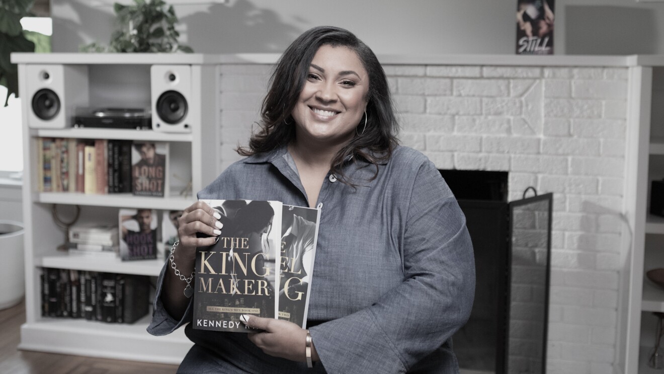 A woman sits in a living room setting, holding two books she wrote and self-published. 