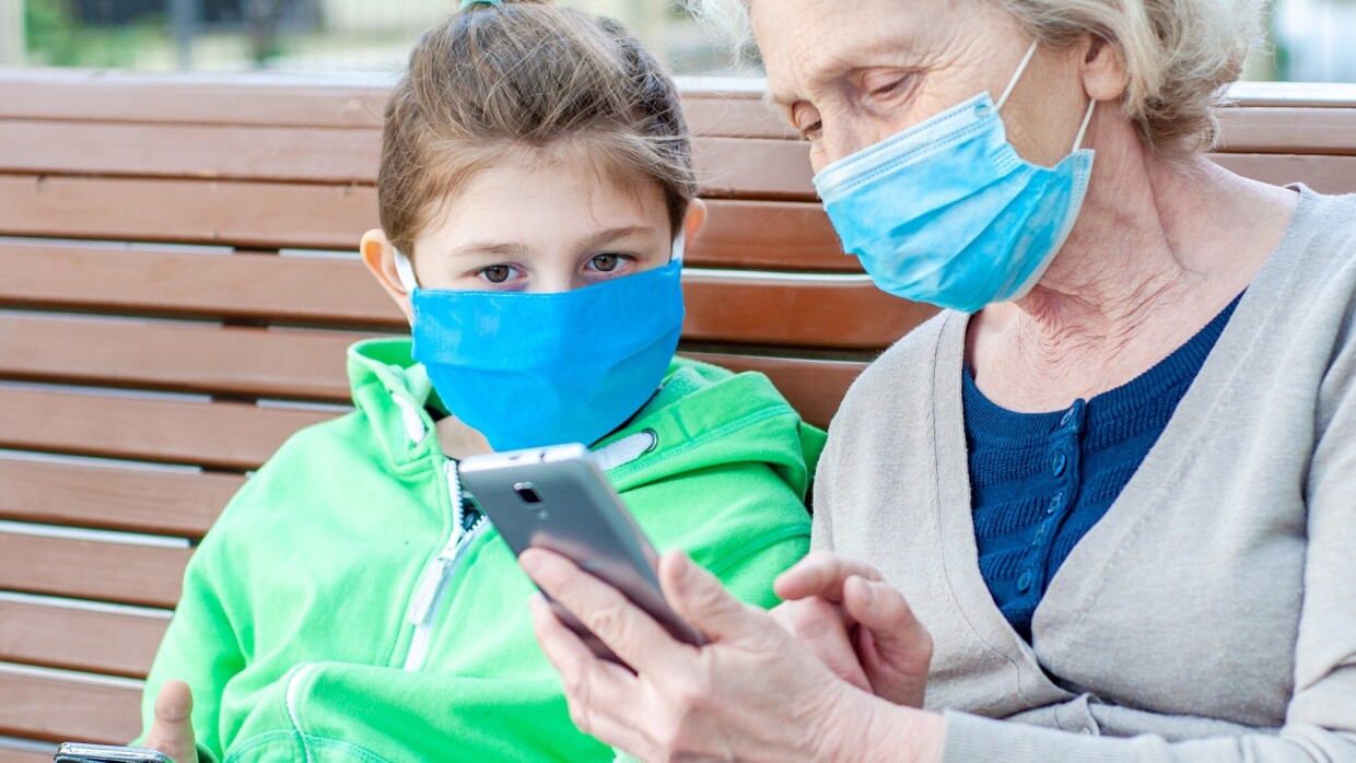 A grandmother sits with her granddaughter on a bench. They are both wearing masks and looking at the grandmother's phone.