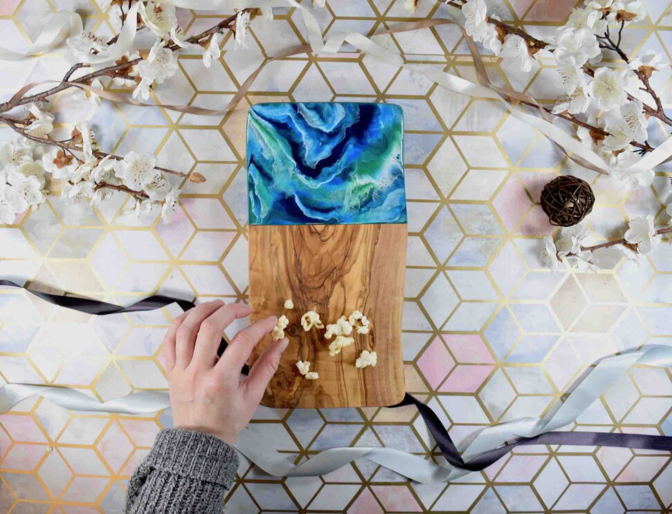 Birds-eye view of someone picking up popcorn from a Kate Chesters Resin Board, There are decorative flowers and ribbons surrounding the board.
