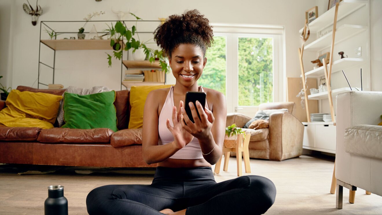 A woman sits on the floor in her living room and plays on her phone. Shis weraing workout clothing and is smiling.