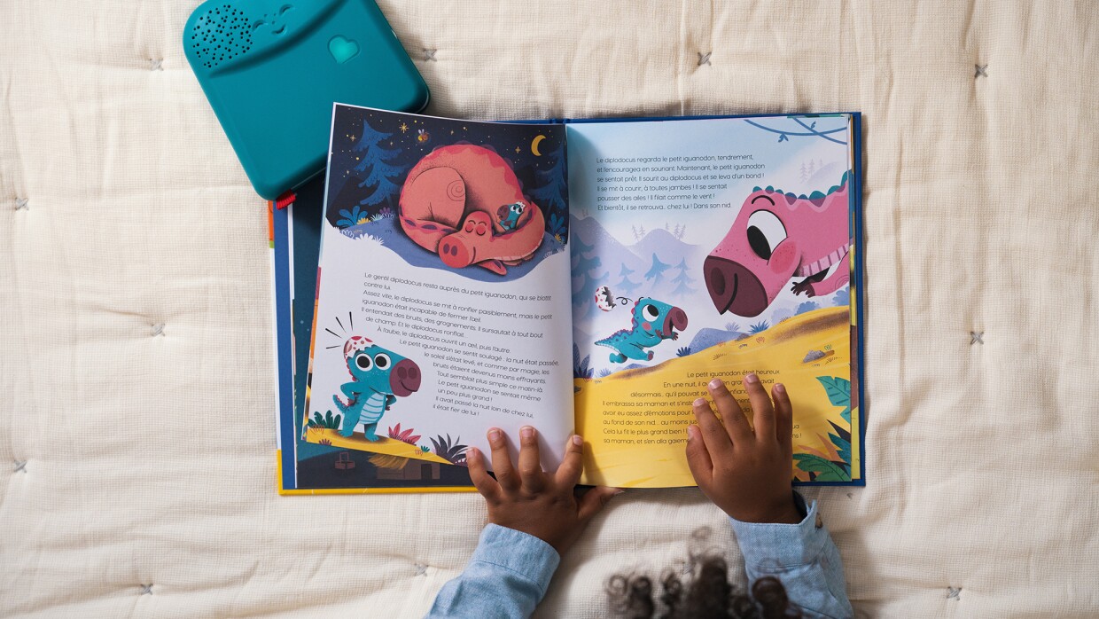 Young child turning pages on a picture book. A device with speaker is near the book and interacting with it. 