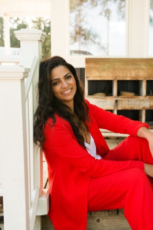 Camila Alves McConaughey wears a red suit and sits on the floor next to a white stair rail 