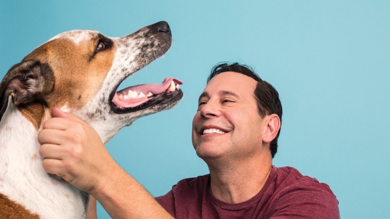 An image of a man smiling while looking at and petting his dog. It is a professional photo against a blue background.