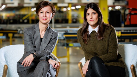 Mariangela Marseglia, vicepresidenta y es la directora general para Italia y España, y Geraldine Leiros, directora general del centro logístico robotizado que Amazon tiene en Illescas (Toledo). Las dos mujeres están sentadas en unos sillones blancos, con las piernas cruzadas y delante de un cinta transportadora de un centro logísticos. Mariangela lleva el pelo largo y un traje chaqueta gris de raya y corte diplomático con una camiseta negra debajo. Lleva anillos en las dos manos, un reloj en la muñeca izquierda y tienes los brazos cruzados y apoyados sobre sus piernas también cruzadas. Lleva el pelo corto de color castaño. Geraldine lleva unos pantalones negros, una camisa blanca y un jersey verde militar de lana. Tiene los brazos apoyados sobre las piernas y el pelo castaño y largo hasta el pecho.